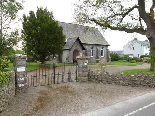 Short stone building with tall roof and tall entrance porch set within large garden grounds. 