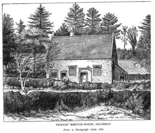 Simple brick building with wooden porch to entrance, situated within heavily wooded area and fields.