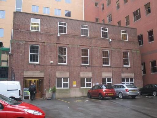 Three storey redbrick building within large car park, surrounded by high-rise structures. 