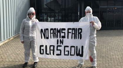 two people in 'blood' spattered hazmat suits holding a banner saying 'no arms fair in Glasgow'