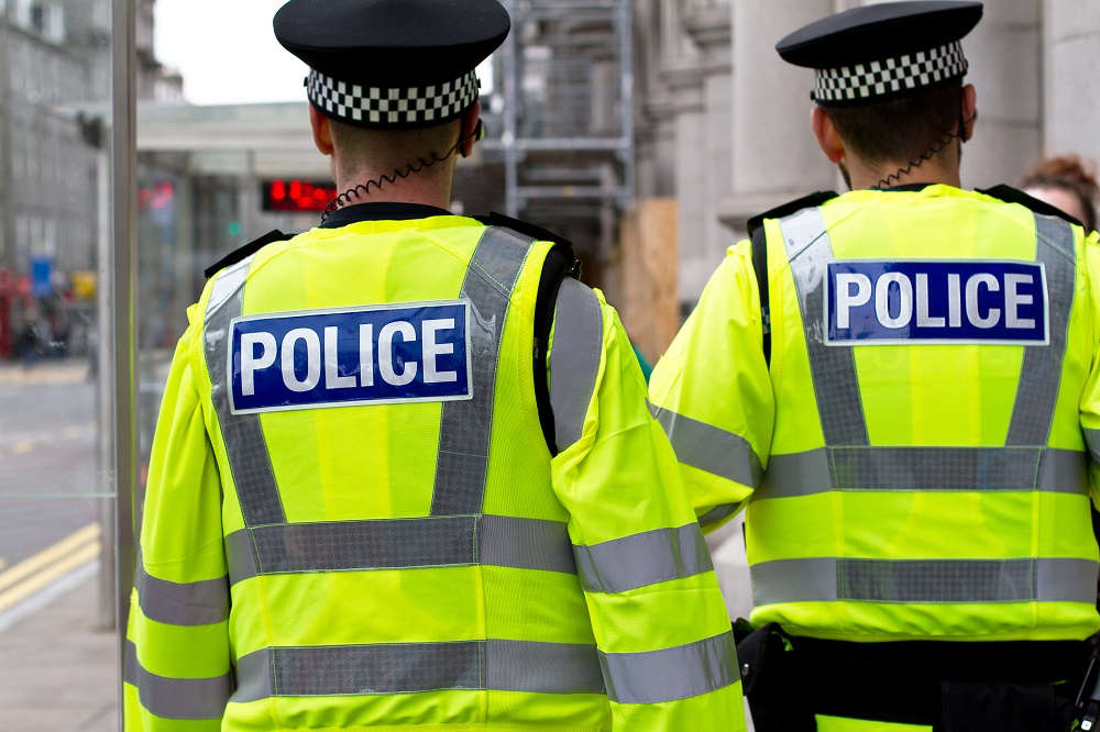 Back of two police officers wearing high-vis police jackets with POLICE written on the back in an urban area