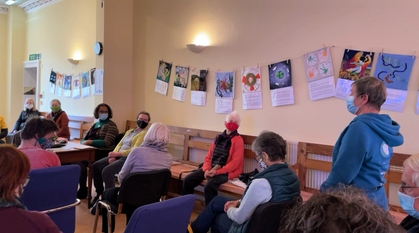 A group of people wearing face masks siting in a circle. One person is standing to give mnistry 