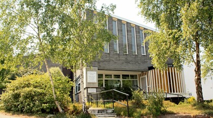 A slate grey, modernist Quaker meeting house with lots of windows and a slanty roof photographed on a sunny day with warm sunnlight dappling through the trees outside.