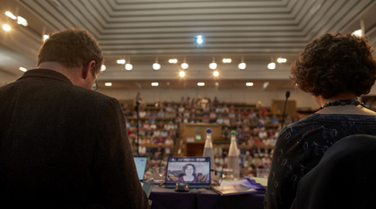 two yearly meeting clerks looking out over face in the large meeting house as one clerk appears on screen with a backdrop of the large meeting house