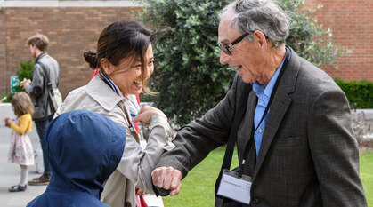 In a garden two adult Quakers do a Covid elbow bump greeting and a child in a hooded jacket  