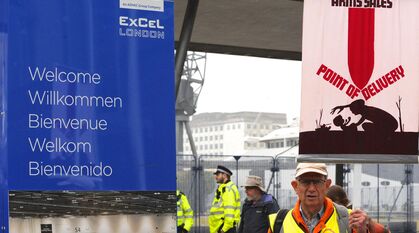 Next to a 'Welcome to the ExCel Centre' sign, a Quaker stands with a banner saying 'arms sales -> point of delivery'. The image on the banner is a simple silhouette drawing of a parent and baby with a rocket coming down on them