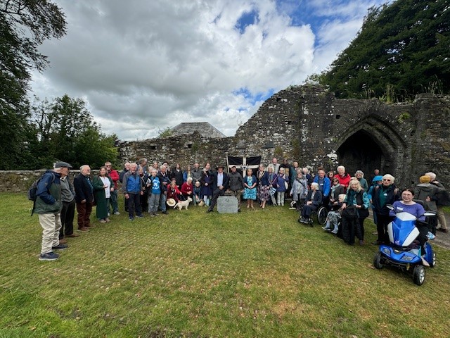 About sixty people gathered around a stone plaque on grass field in front of old stone building ruins.  