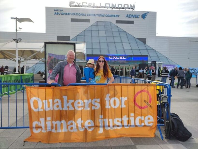 Two adults and a child standing by a banner outside the ExCel Centre in London. The banner reads 'Quakers for Climate Justice' 