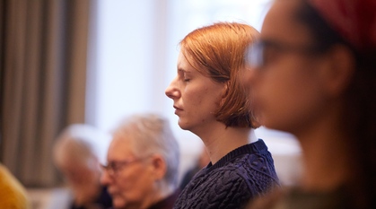 Close up on one person in meeting for worship. The image is soft and both the main subject and others around her have their eyes closed, listening for the small, still voice