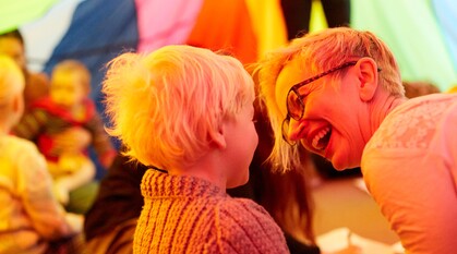 At an all-age event under a parachute, a person laughs with a child