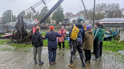 A group of people stand, mostly listening and one speaking, by the docks