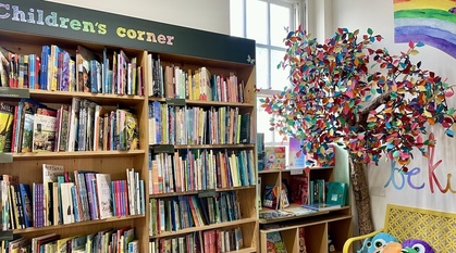 The children's corner at the Quaker Bookshop with shelves of exciting children's books, a tree made of papier maché, a rainbow and paper flowers on the walls and a child height bench in yellow with fun, colourful cushions on it