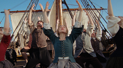 A group of people in historical clothes, with a woman in the centre. They are dancing, with their arms in the air. In the background the rigging of a ship is visible.