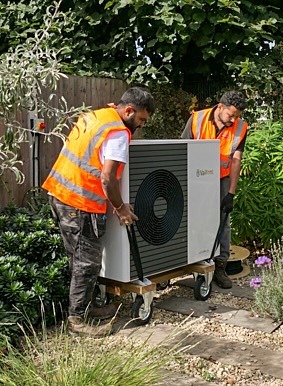 Two installers moving a heat pump through a garden outside an Edwardian house. Photo: Katy King / Nesta / Climate Visuals