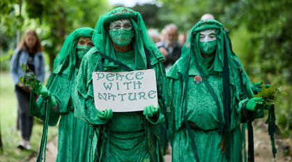 A group of people dressed all in green walk towards the camera,  through a park. The lead person carries a sign saying 'peace with nature'.