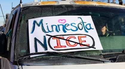 close-up of a dirty van covered with a a placard attached to the windscreen which says Minnesota No ICE with a heart over Minnesota. Part of a woman's face is visible from the outside to the driver's seat