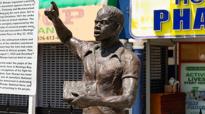 A photograph of a statue of Sam Sharpe designed by Kay Sullivan, in Montego Bay, Jamaica; it's a sculpture of a Black man with his arm raised, holding a book in the other hand, and speaking